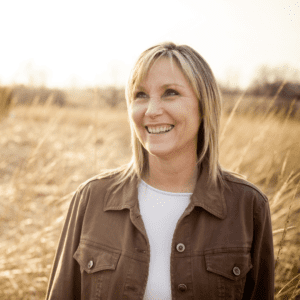 Blonde lady standing in a field smiling with brown jacket on